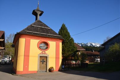 Ein kleines, buntes Gebäude mit einem runden Fenster und einem spitzen Dach. Im Hintergrund sind schneebedeckte Berge und Bäume zu sehen.
