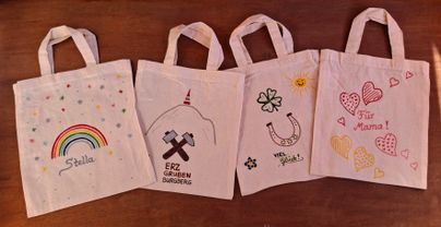 Vier bunte Tragetaschen mit verschiedenen Designs liegen auf einem Tisch. Jede Tasche hat ein einzigartiges Motiv, darunter ein Regenbogen und Herzen.