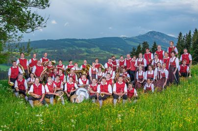 Eine große Gruppe von Musikern steht in einer blühenden Wiese mit Bergen im Hintergrund. Sie tragen traditionelle Kleidung in Rot und Weiß und halten verschiedene Instrumente.