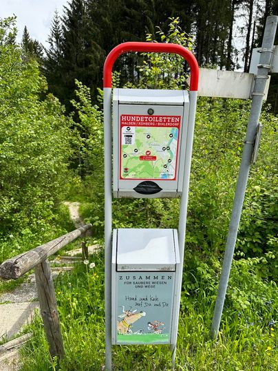 Ein Informationstafel für Hundetoiletten steht inmitten von Bäumen und Grün. Darunter befindet sich ein Behälter für Hundekot.