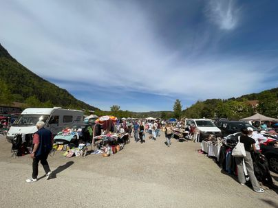 Ein belebter Flohmarkt mit verschiedenen Ständen und Menschen. Im Hintergrund sind grüne Hügel und ein blauer Himmel zu sehen.