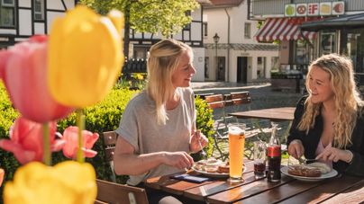 Zwei Frauen sitzen draußen an einem Tisch und genießen ihr Essen. Im Vordergrund sind bunte Tulpen zu sehen.
