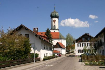 Eine ruhige Straße in einem bayerischen Dorf mit traditionellen Häusern und einer markanten Kirche im Hintergrund. Der Himmel ist klar und blau, was eine friedliche Atmosphäre schafft.