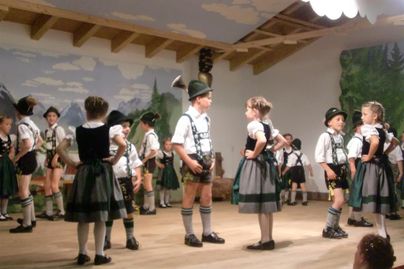 Eine Gruppe von Kindern in traditionellen Trachten tanzt auf einer Bühne. Die Kulisse zeigt Berge und eine ländliche Landschaft.