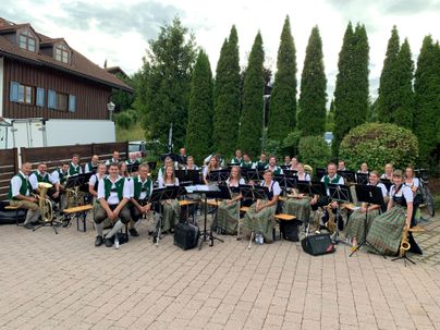 Eine Musikkapelle posiert im Freien, umgeben von hohen Tannen und einem Gebäude im Hintergrund. Die Mitglieder tragen traditionelle Kleidung und sitzen mit ihren Instrumenten.