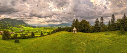 Eine weitläufige grüne Landschaft mit sanften Hügeln und einem Pavillon. Im Hintergrund sind Berge und eine bewölkte Himmel zu sehen.