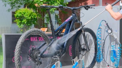 Ein Fahrrad wird gründlich gereinigt. Im Hintergrund sind grüne Bäume und eine Fahrradwerkstatt zu sehen.