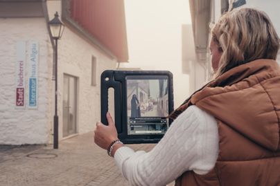 Eine Person hält ein digitales Gerät und betrachtet eine Straße. Im Hintergrund sind historische Gebäude zu sehen.