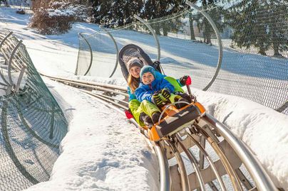 Zwei Kindern rodeln mit dem Alpsee Coaster der Alpsee Bergwelt bei Immenstadt im Winter den Berg hinunter.
