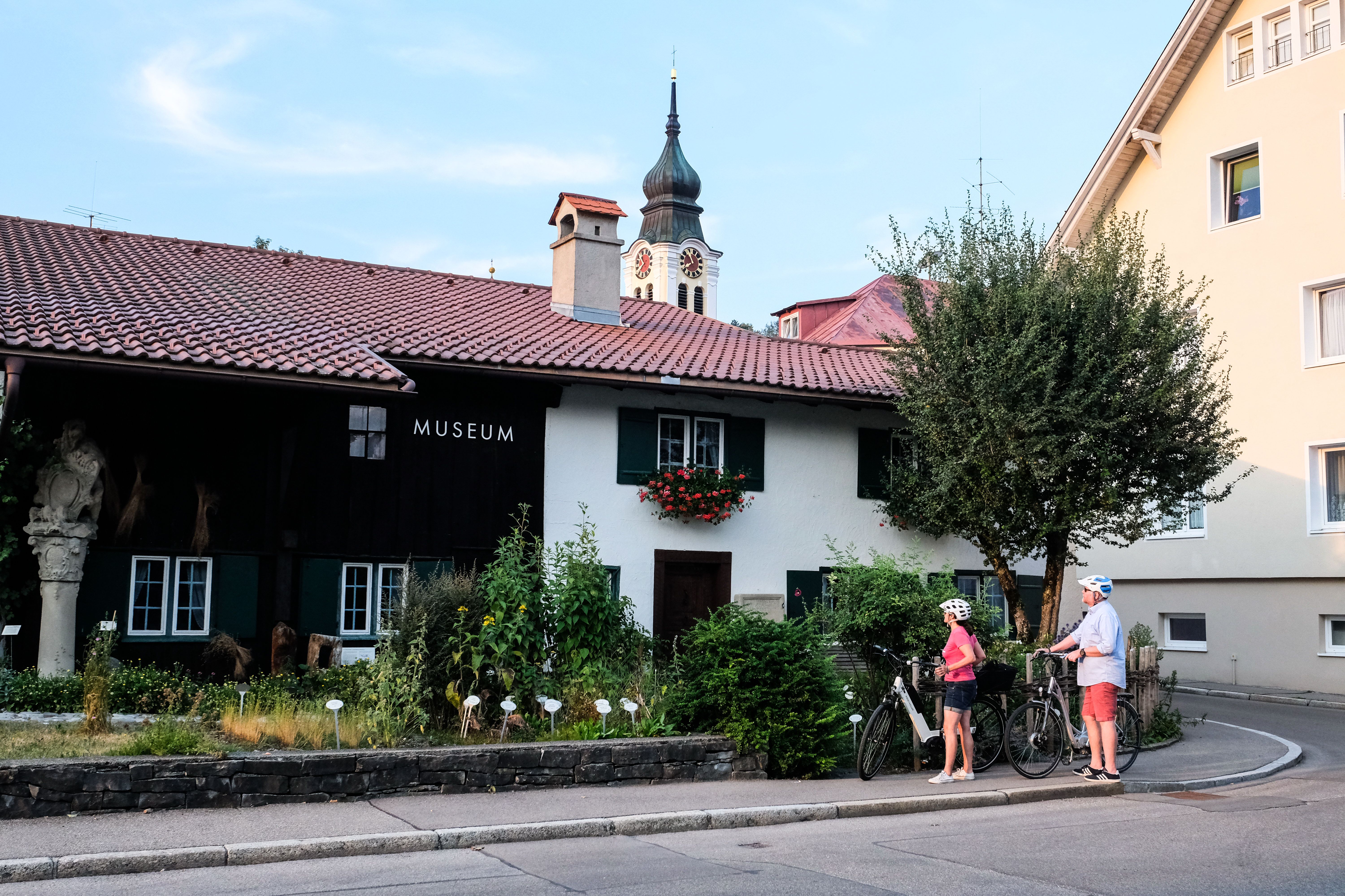 Zwei Fahrradfahrer bewundern das Heimathaus Museum in Sonthofen, im Hintergrund sieht man den Kirchturm der St. Michael Kirche. Das Heimathaus Sonthofen ist in einem historischen Allgäuer Bauernhaus aus dem 18. Jahrhundert untergebracht und befindet sich im ältesten Teil von Sonthofen.