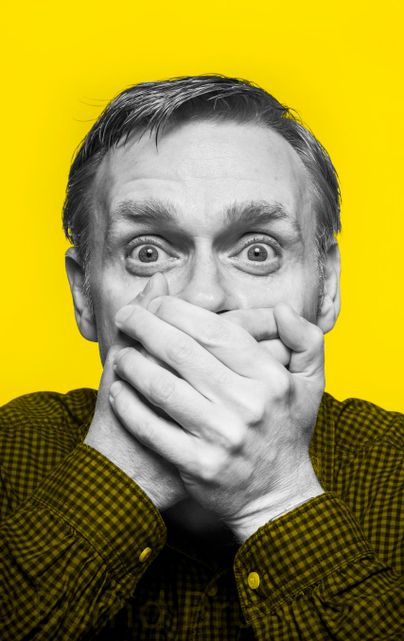 Ein Mann mit einem erstaunten Gesichtsausdruck hält sich die Hände vor den Mund. Der Hintergrund ist leuchtend gelb, was die Emotion verstärkt.