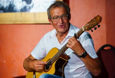 Ein Mann spielt Gitarre und lächelt dabei. Der Hintergrund ist rot und es gibt keine weiteren Ablenkungen.