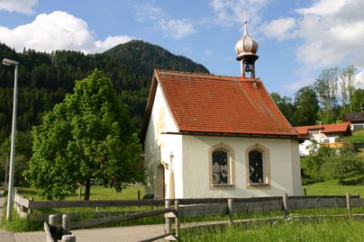 Eine kleine, charmante Kapelle steht in einer malerischen Landschaft. Im Hintergrund sind grüne Bäume und Berge zu sehen.
