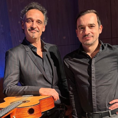 Zwei Musiker stehen nebeneinander auf der Bühne. Einer hält eine Gitarre und beide tragen elegante schwarze Kleidung.