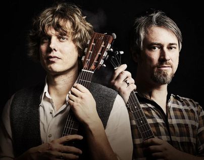 Zwei Musiker stehen nebeneinander, jeder mit einer Gitarre in der Hand. Der Hintergrund ist dunkel, was die Gesichter und Instrumente hervorhebt.