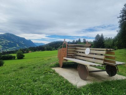 Eine kreative Bank steht auf einer Wiese mit Blick auf die Berge. Der Himmel ist leicht bewölkt und die Landschaft ist grün und einladend.