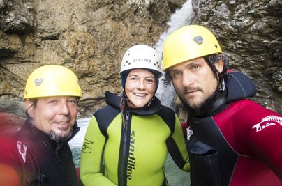 Drei Personen in Neoprenanzügen und Helmen stehen vor einem Wasserfall. Sie lächeln und scheinen bereit für ein Abenteuer im Wasser.
