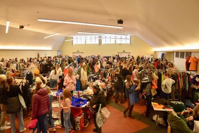 Ein belebter Secondhand-Markt mit vielen Menschen, die Kleidung durchsuchen. Zahlreiche Stände und Regale sind mit Kleidung und Accessoires gefüllt.