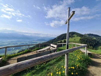 Eine ruhige Berglandschaft mit einem Holzkreuz und einer schönen Aussicht. Die Wiesen sind grün und mit gelben Blumen bedeckt.