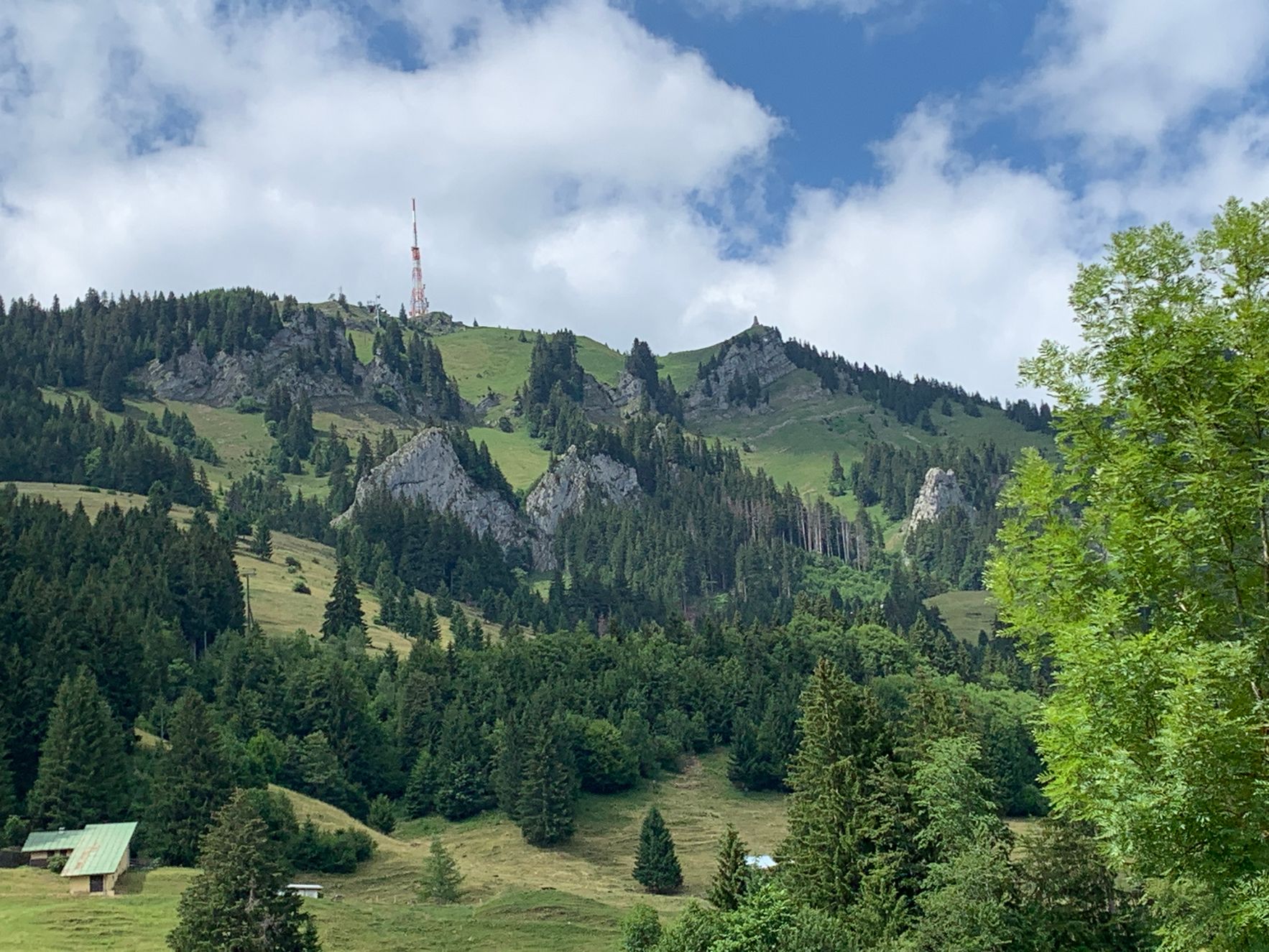 Die Aufnahme zeigt eine Berglandschaft mit einem Senderturm und vielen Bäumen. Es ist tagsüber und die Wolken sind sichtbar.