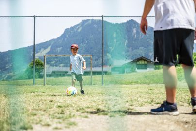 Ein Kind spielt Fußball auf einem Platz mit einem Tor im Hintergrund. Die Berge sind klar sichtbar und die Sonne scheint.