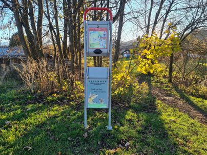 Ein Informationsschild steht auf einem grünen Weg, umgeben von Bäumen und Sträuchern. Es zeigt eine Karte und bietet Informationen für Spaziergänger.