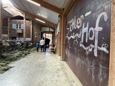 Ein Pferdehof mit Tieren und einer großen Wandtafel. Menschen gehen einen Stallgang entlang, umgeben von Heu und Geräten.