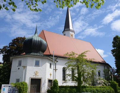 Eine beeindruckende Kirche mit einem spitzen Turm und einem markanten Dach. Umgeben von grünem Laub und einem klaren blauen Himmel.