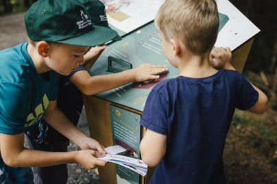 Zwei Kinder erkunden ein interaktives Infoterminal im Freien. Der Junge in blauem T-Shirt hält ein Blatt in der Hand, während der andere auf die Oberfläche zeigt.