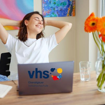 Eine entspannte junge Frau sitzt am Schreibtisch und genießt ihre Zeit. Vor ihr steht ein Laptop mit dem Logo einer Volkshochschule.