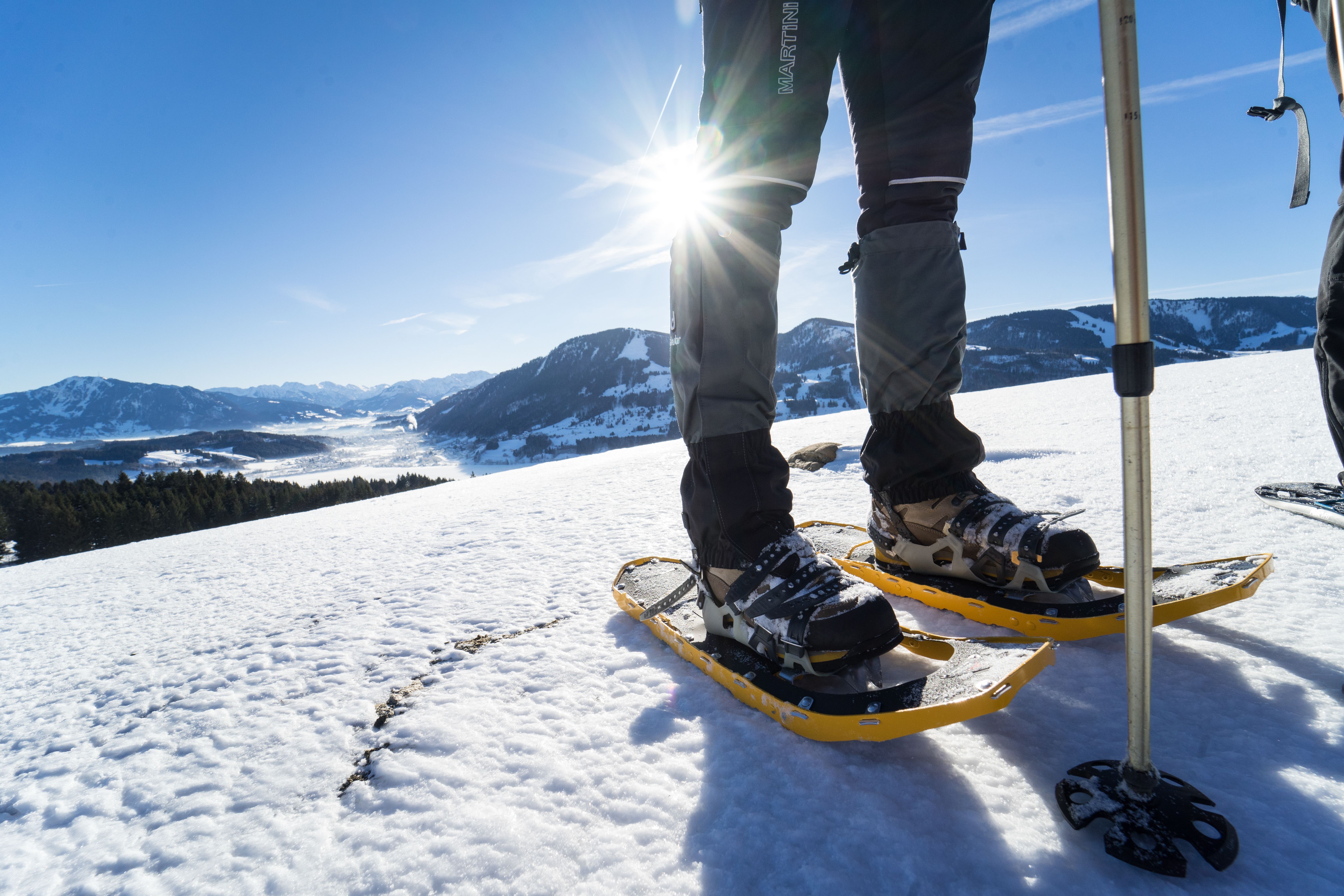 Ein Wanderer steht mit seinen gelben Schneeschuhen im Winter auf einer Anhöhe über Immenstadt. Zum Schneeschuhlaufen braucht es nicht viel. Warme, hochgeschnittene Wanderschuhe und ein paar Teleskopstöcke mit großen Tiefschneetellern.