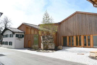 Ein modernes Gebäude mit Holzfassade und großen Fenstern steht neben einem traditionellen Haus. Vor dem Gebäude befindet sich ein großer Stein und ein Baum.