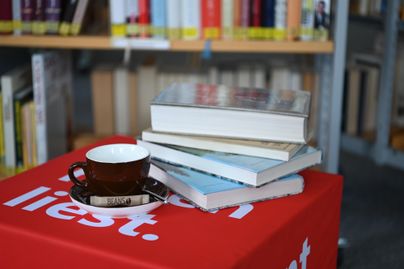 Ein Stapel Bücher liegt auf einem Tisch mit einer Tasse Kaffee und einer Untertasse. Im Hintergrund sind Regale mit weiteren Büchern zu sehen.