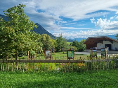 Eine grüne Landschaft mit Bäumen und einem kleinen Gebäude im Hintergrund. Im Vordergrund sind einige Informationstafeln aufgestellt.