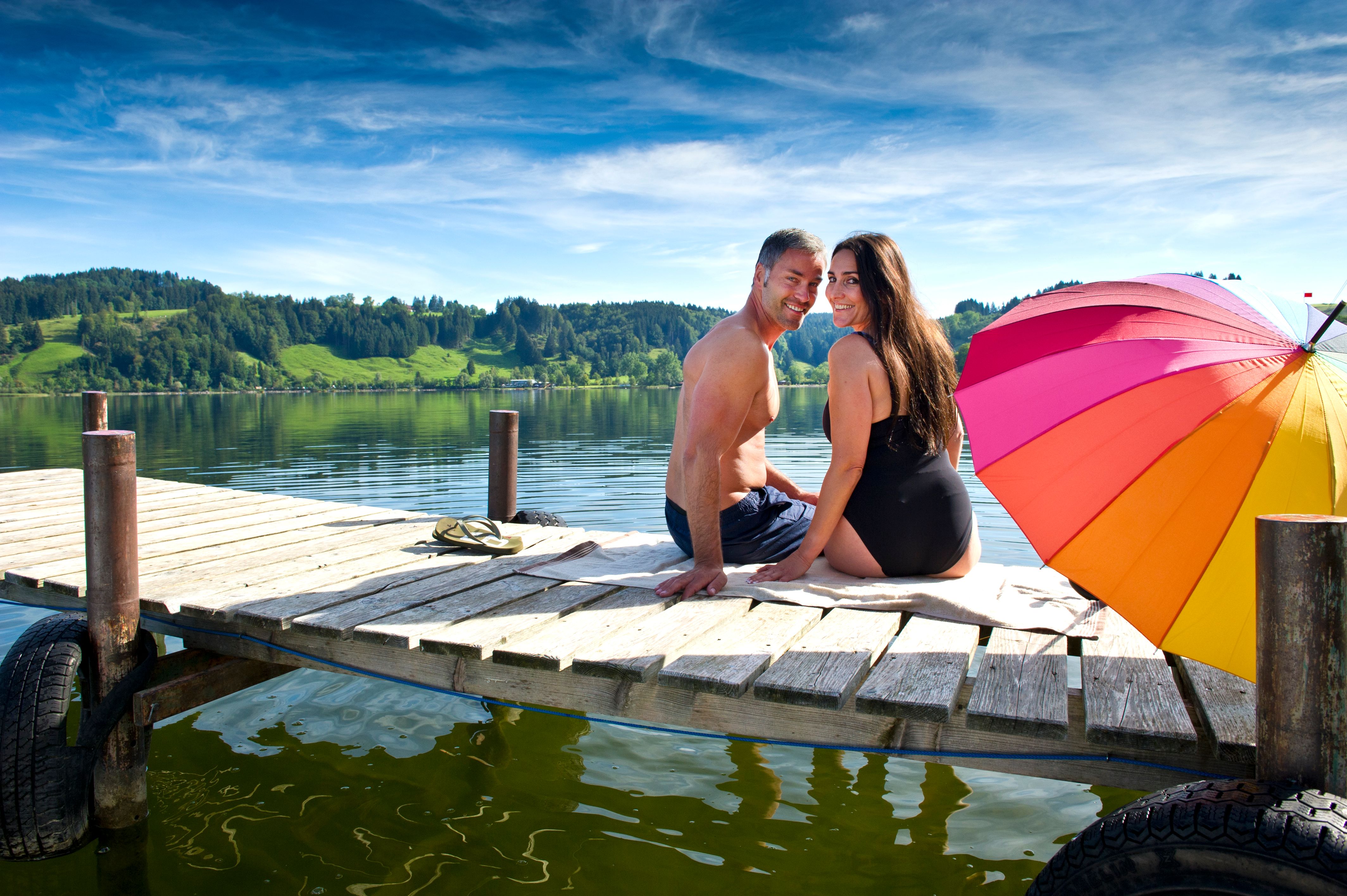 Ein Paar sitzt in Badekleidung auf dem Steg am Großen Alpsee in Immenstadt mit einem Sonnenschirm.