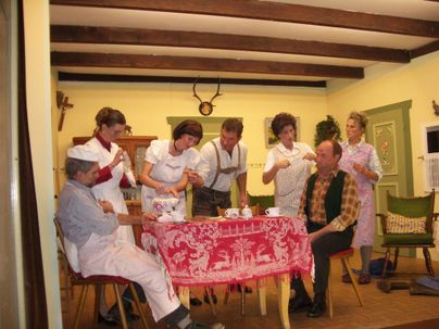 Eine Gruppe von Menschen sitzt um einen Tisch in einem gemütlichen Raum. Sie tragen traditionelle Kleidung und scheinen an einer geselligen Teestunde teilzunehmen.