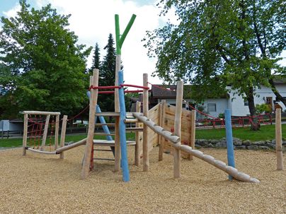 Ein Spielplatz aus Holz mit verschiedenen Kletterelementen und Rutschen. Der Boden ist mit feinem Kies bedeckt und es gibt viele Bäume im Hintergrund.