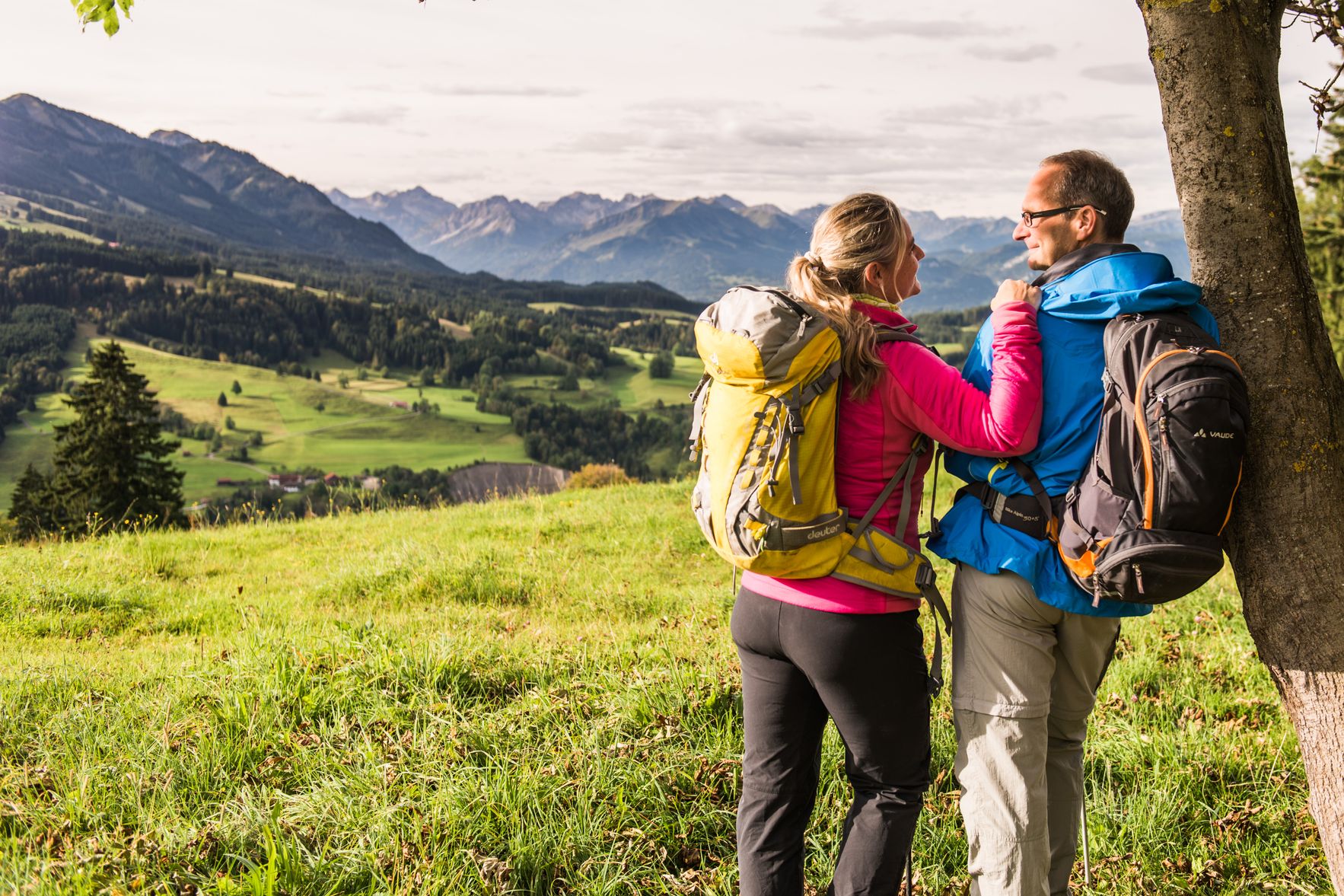 Zwei Wanderer lehnen in Wanderkleidung und mit Rucksäcken an einen Baum in Sonthofen.
