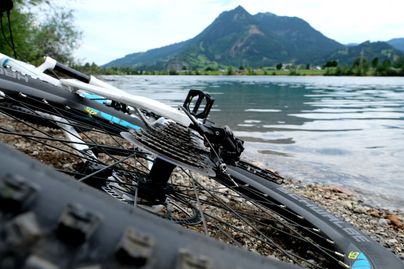 Ein weißes Fahrrad liegt am Ufer eines Sees. Im Hintergrund liegt der Grünten.