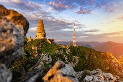 Ein Aussichtspunkt mit einem historischen Turm auf einem Berg. Im Hintergrund sind sanfte Hügel und ein farbenfroher Himmel zu sehen.