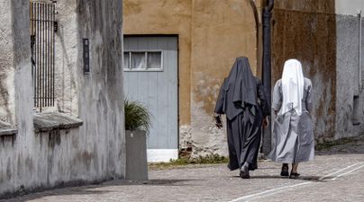 Zwei Nonnen gehen eine Geschäftsstraße entlang. Im Hintergrund ist eine alte Wand und eine graue Tür zu sehen.