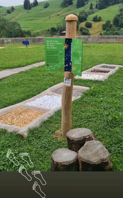 Ein Barfußweg in einer grünen Wiese, umgeben von Hügeln. Neben dem Weg steht ein Informationsschild.