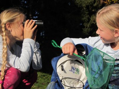 Zwei Kinder spielen im Freien. Eines schaut durch ein Fernglas, während das andere eine Fangnetz aus einem Rucksack holt.