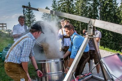Eine Gruppe von Männern in traditionellen Trachten arbeitet zusammen an einem großen Behälter, aus dem Dampf aufsteigt. Sie befinden sich im Freien, umgeben von Bäumen und einer schönen Landschaft.