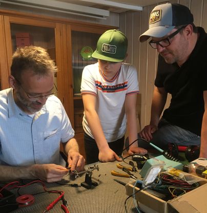 Drei Personen arbeiten zusammen an einem Tisch mit Werkzeugen und elektrischen Komponenten. Sie zeigen Interesse an den Bastelarbeiten und lernen voneinander.