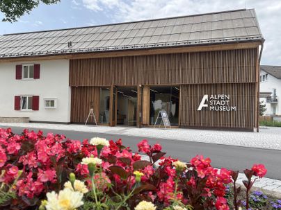 Ein modernes Gebäude mit Holzfassade beherbergt das Alpenstadt Museum. Im Vordergrund blühen bunte Pflanzen.