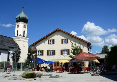 Ein malerischer Platz mit einem Restaurant und bunten Sonnenschirmen. Im Hintergrund ist ein Kirchturm und blauer Himmel mit einigen Wolken zu sehen.