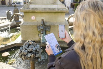 Eine Person steht vor einem Brunnen und hält ein Smartphone sowie einen Flyer in der Hand. Im Hintergrund sind Teile des Brunnens und die Umgebung sichtbar.