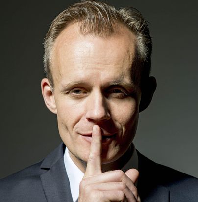 Ein Mann in einem Anzug macht mit dem Finger ein "Still"-Zeichen. Er blickt direkt in die Kamera und hat ein verschmitztes Lächeln.