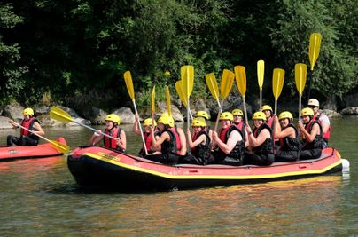 Eine Gruppe von Menschen paddelt in einem Rafting-Boot auf einem ruhigen Fluss. Sie tragen Helme und Schwimmwesten und sind bereit für ein Abenteuer.